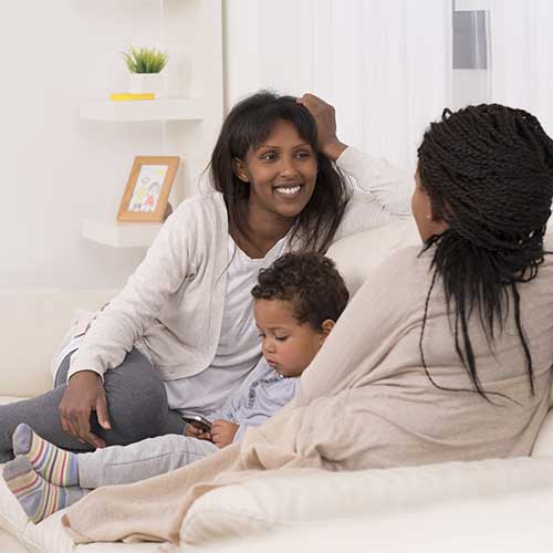 Two women sit on couch talking, with small child sitting between them